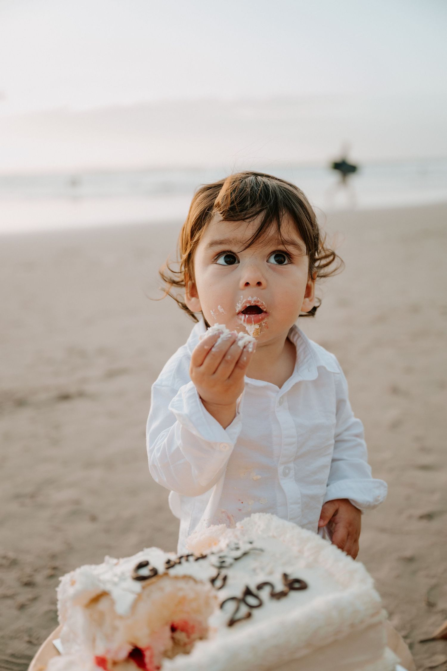 Toddler in white shirt eating birthday cake on beach, with 2026 written on white frosted cake.