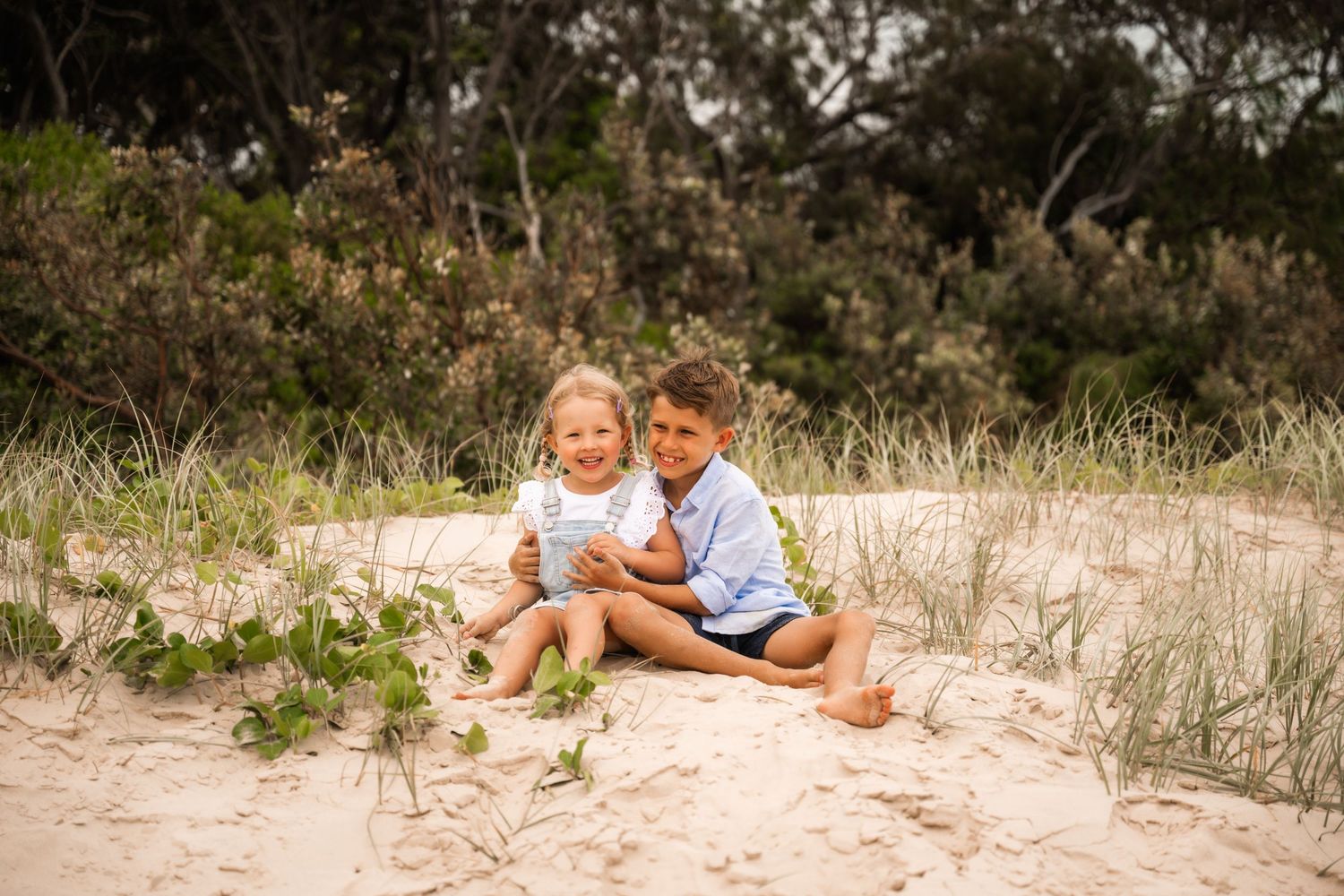 Two happy children sit together on sandy beach dunes surrounded by coastal vegetation in a warm outdoor portrait.