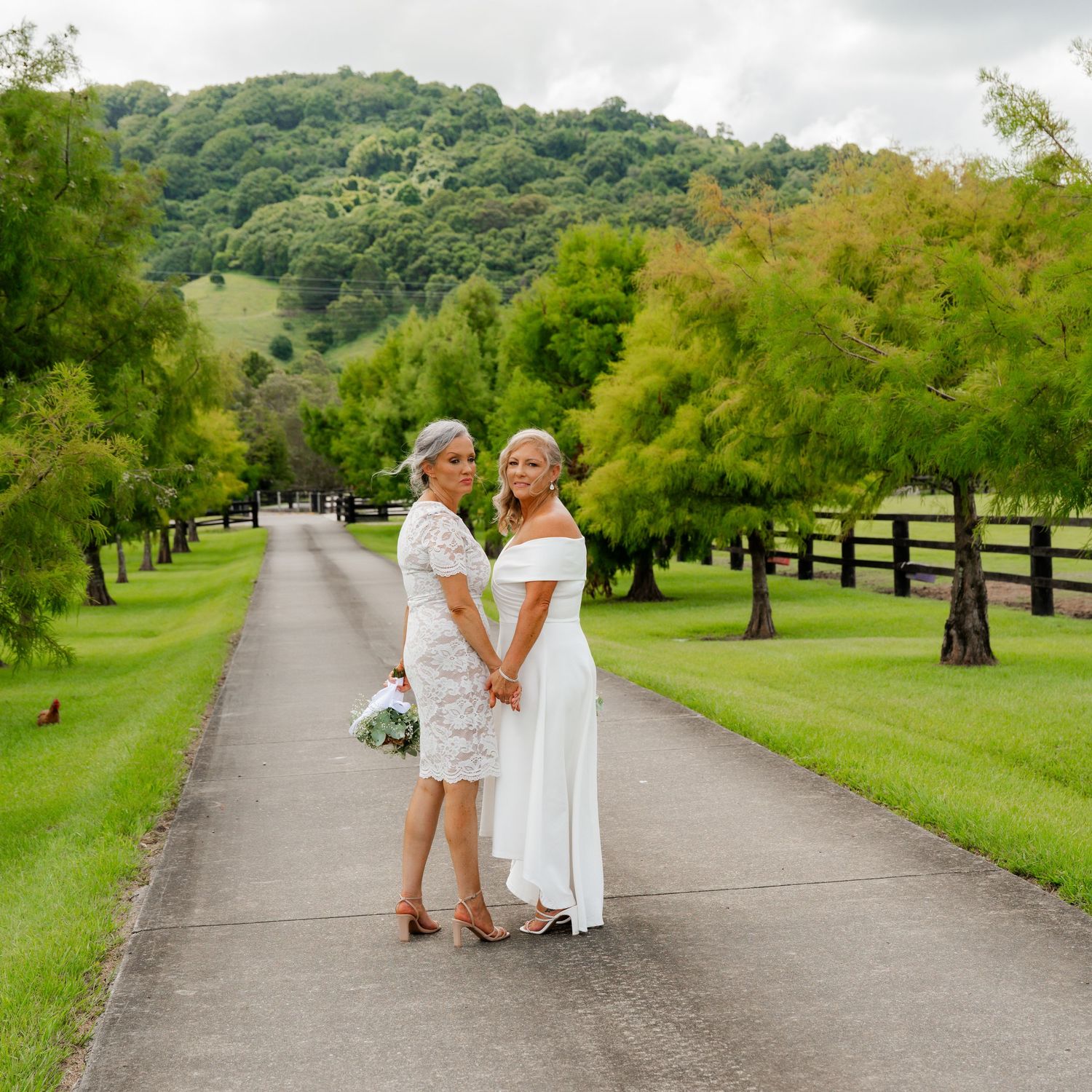 Two brides in white dresses hold hands on a tree-lined driveway with green hills in the background.