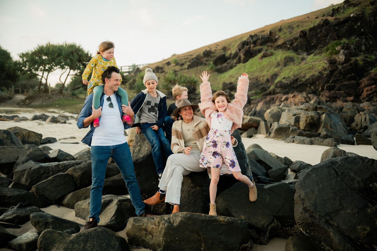 Family of six joyfully posing on rocky beach with green hills, wearing colorful outfits, child on shoulders.