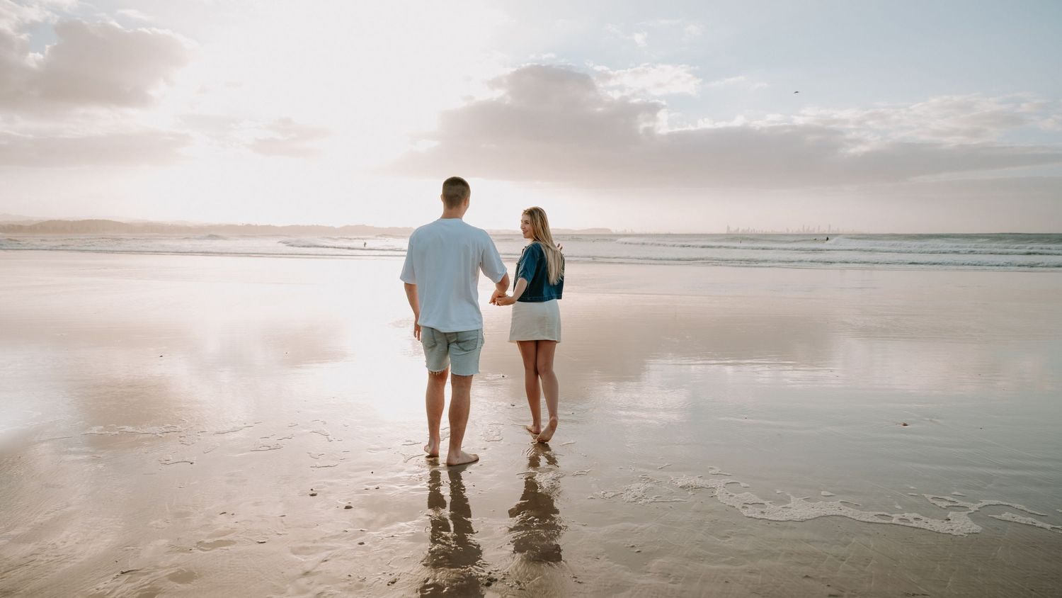 A couple walks hand in hand on a reflective beach at sunset, waves gently lapping the shore.