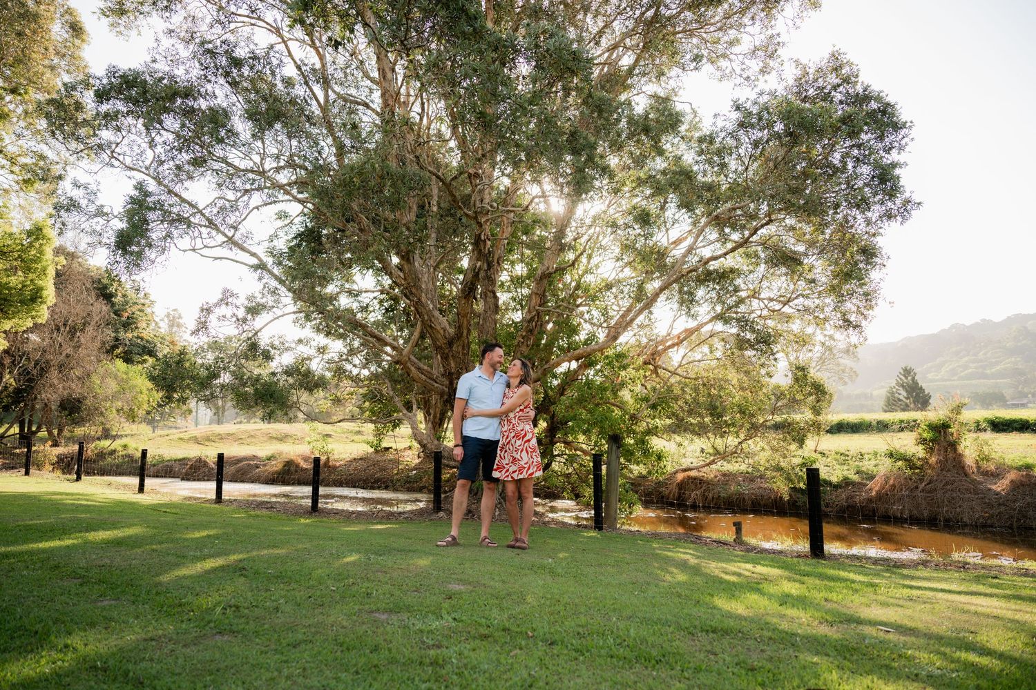 Couple embracing under a large shady tree on a green lawn beside a wooden fence and creek, with hills glowing in warm golden sunlight.