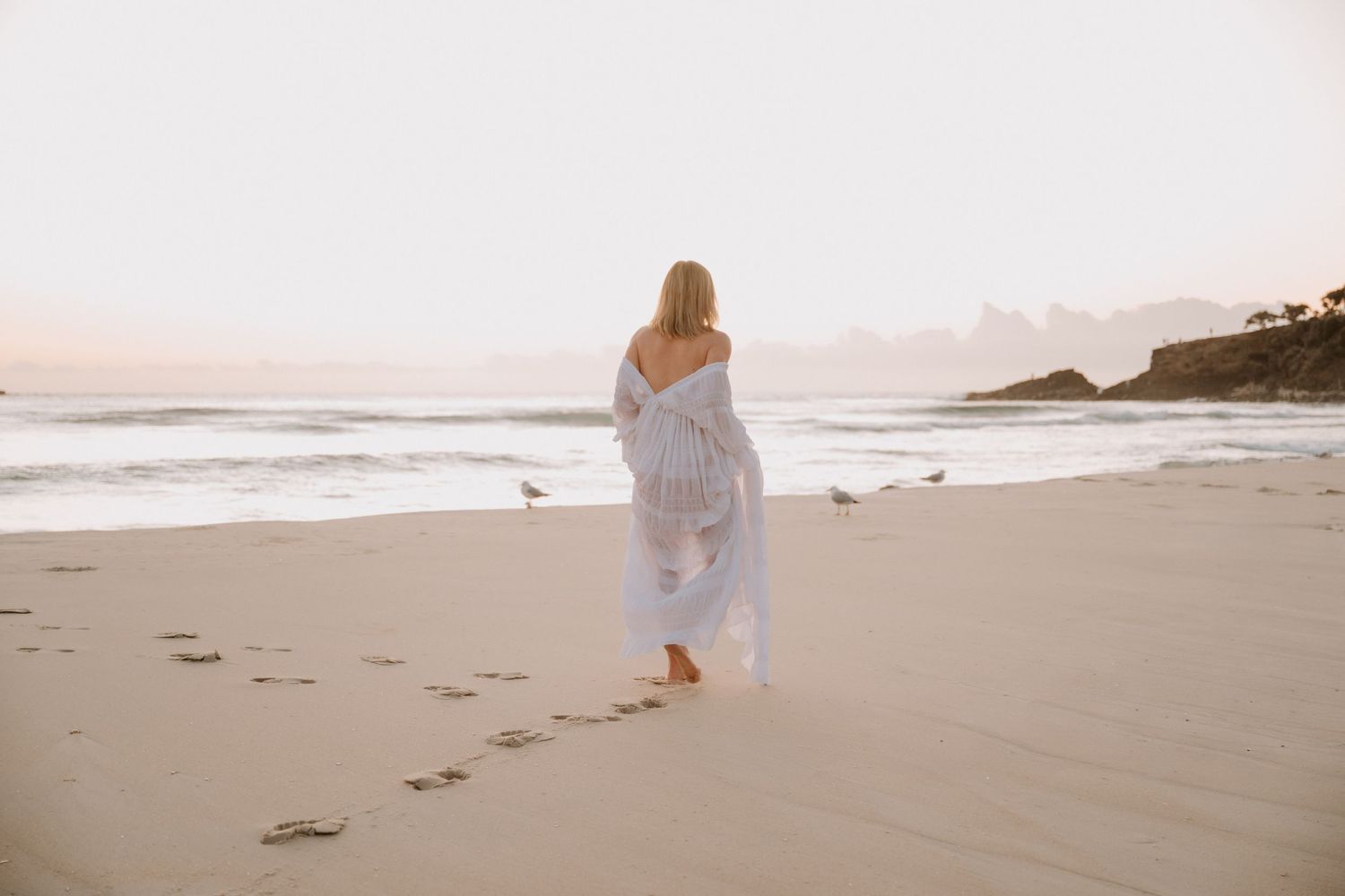 Woman in white dress walking barefoot on sandy beach at sunset, leaving footprints, with ocean waves and seagulls nearby.