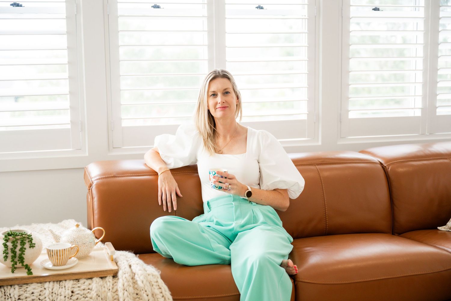 Woman in white top and mint green pants lounges on brown leather couch near bright windows.