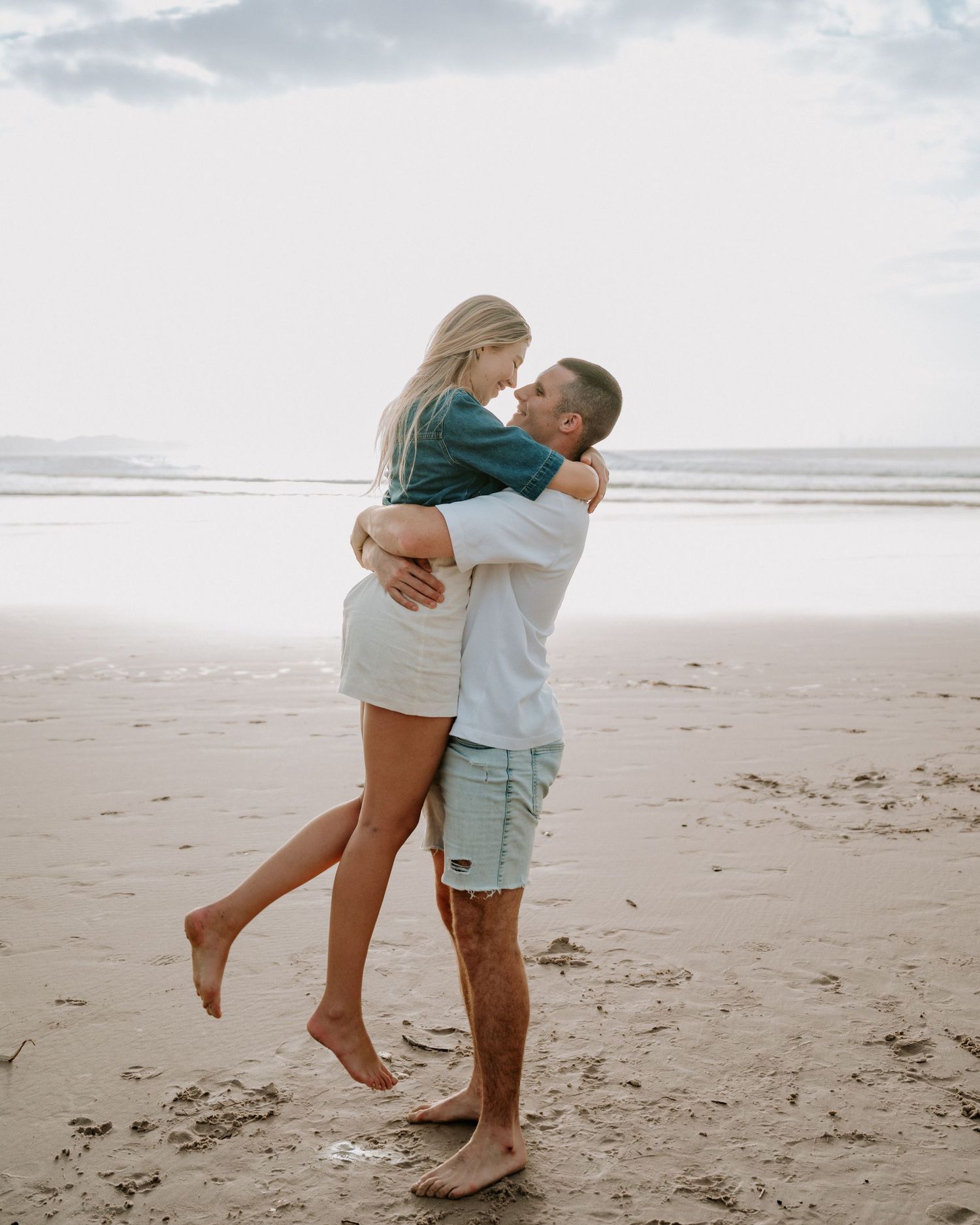 Couple embracing on a sandy beach, man lifting woman as they share a romantic moment with ocean in background.