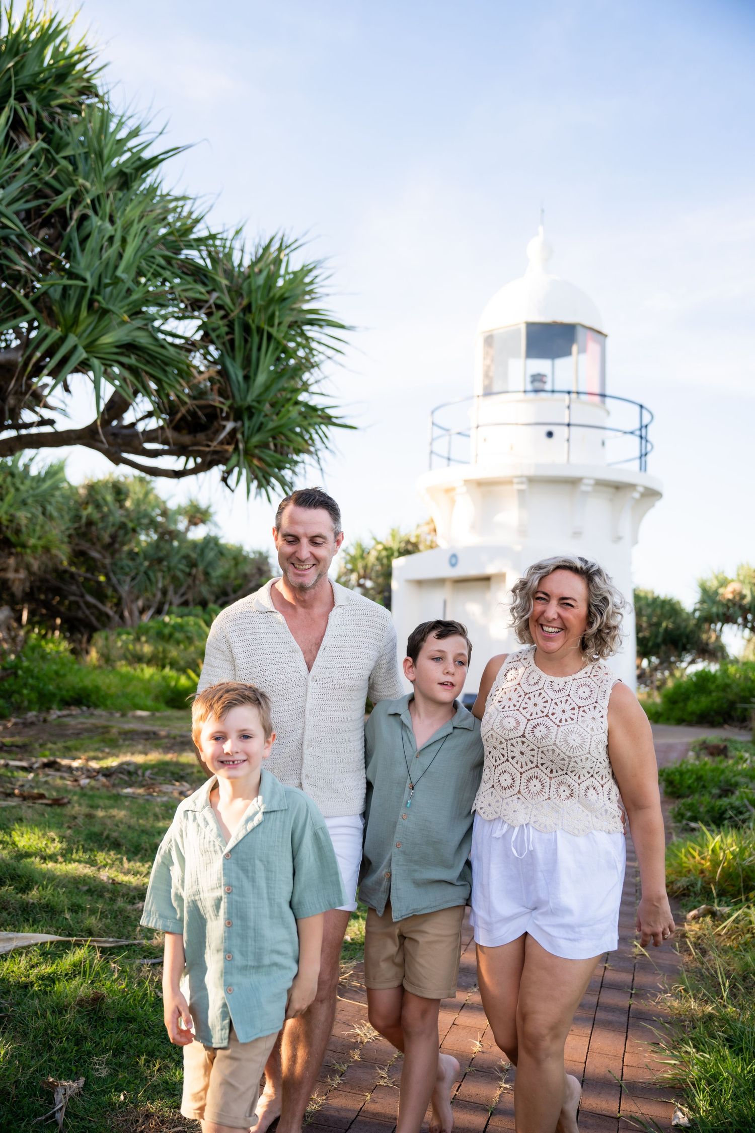 Family of four smiling in front of a white lighthouse surrounded by lush greenery on a sunny day.