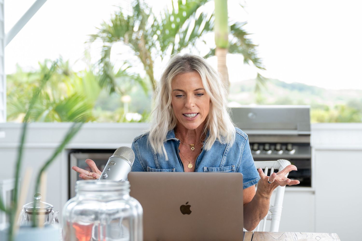 Blonde woman in denim shirt gesturing while video calling on MacBook laptop in bright modern kitchen.