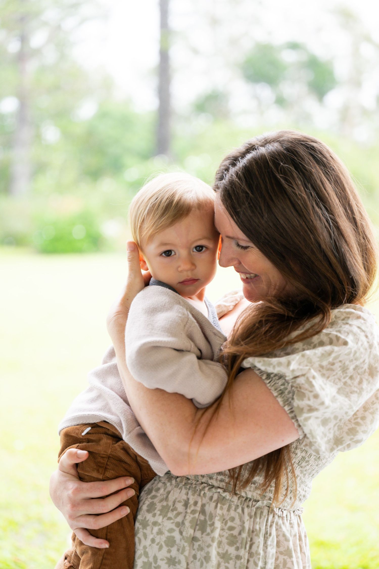 A tender moment captured outdoors as a parent in a floral dress holds a baby wearing white and brown against a soft green background.