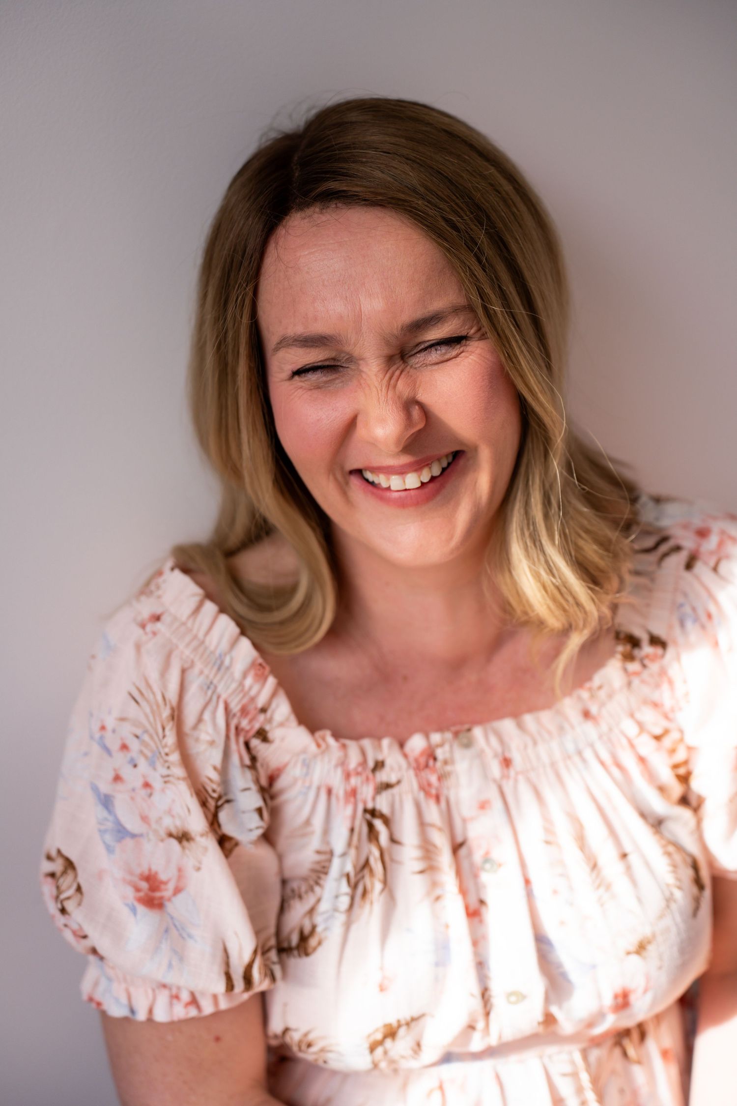 A smiling woman with blonde hair wearing a floral off-shoulder top laughs joyfully against a light gray background.