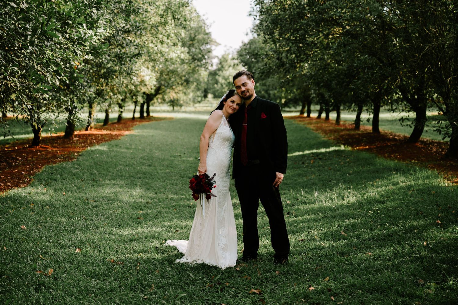 Bride and groom posing together in a lush green orchard aisle, bride holding a deep red bouquet in a white gown.