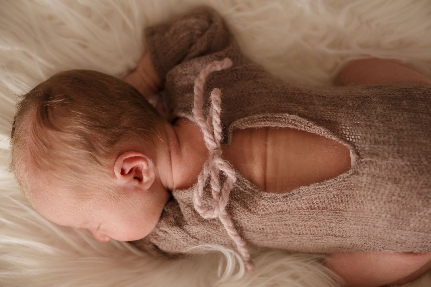Sleeping newborn baby wearing a knit beige romper with bow tie detail, resting on a soft white fur blanket.