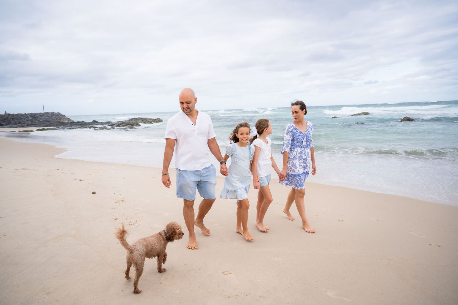 Family with two girls and a dog walking barefoot on a sandy beach with ocean waves in the background.