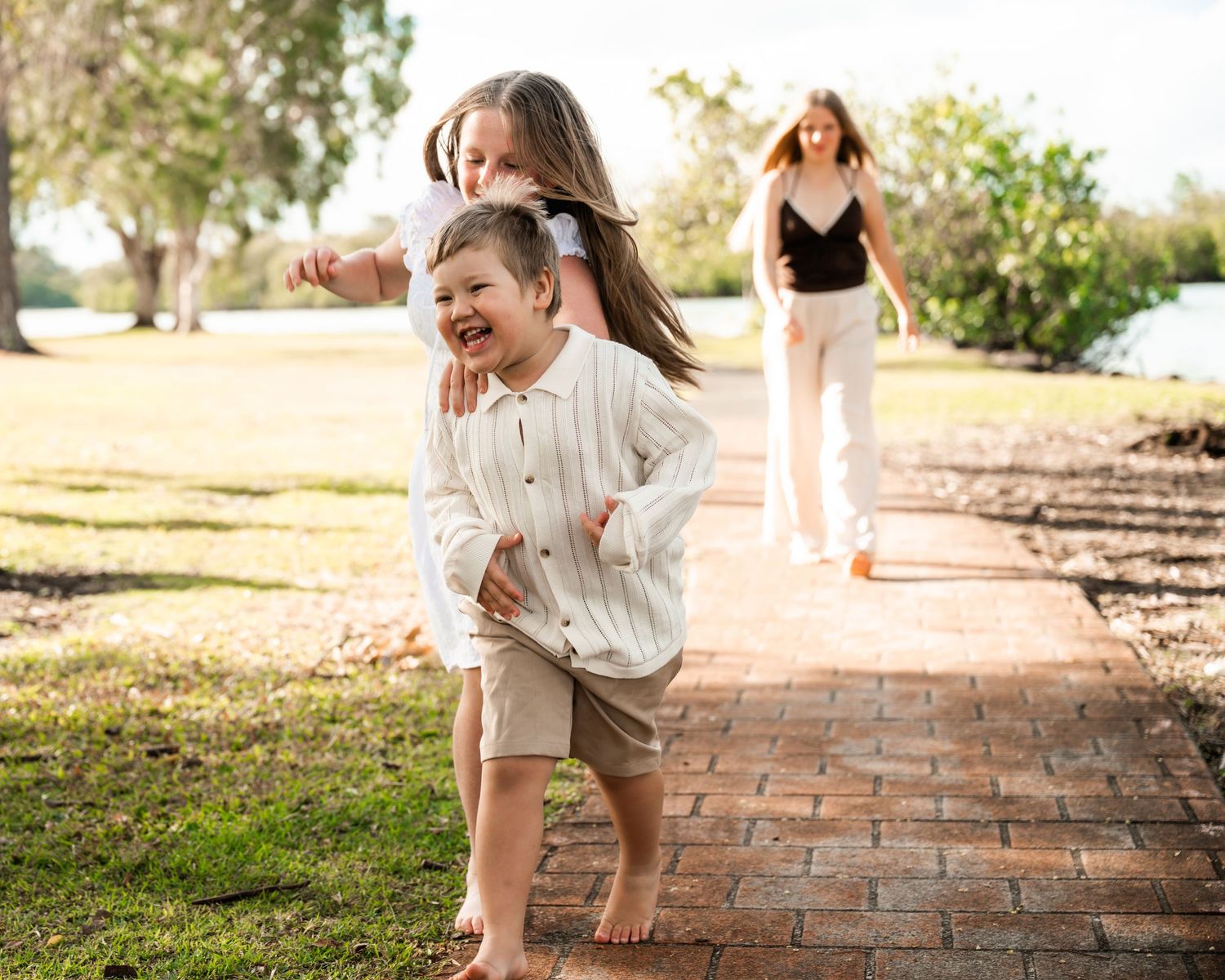Two children playing joyfully on a brick path in a sunny park, with a woman walking behind them.