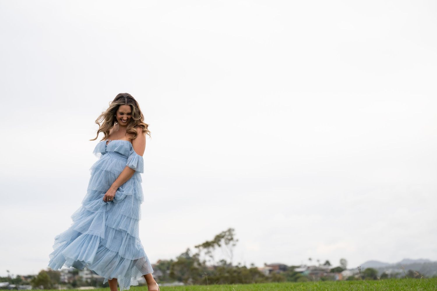 Pregnant woman in a flowing light blue off-shoulder tiered maxi dress walking gracefully on green grass outdoors.