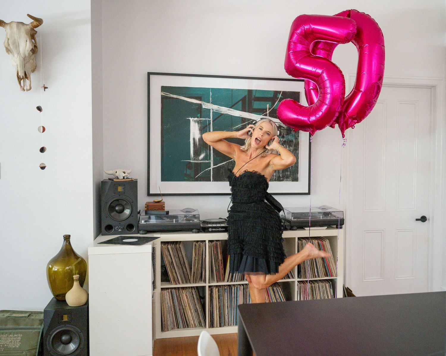 Woman in black dress jumping joyfully holding a pink number 50 balloon in a vinyl record-filled living room.