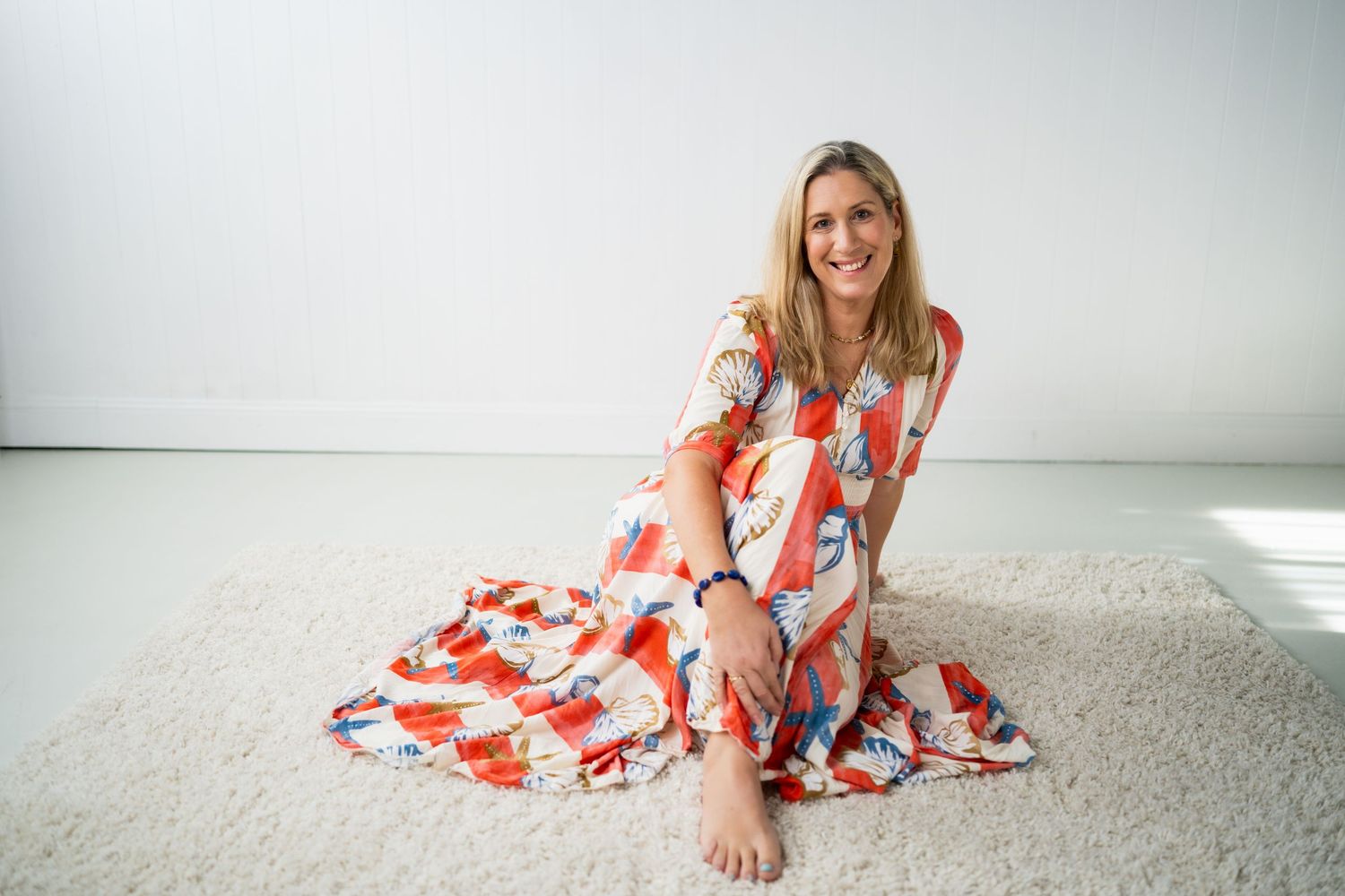 Woman in floral maxi dress sitting barefoot on cream carpet against white wall, smiling warmly.
