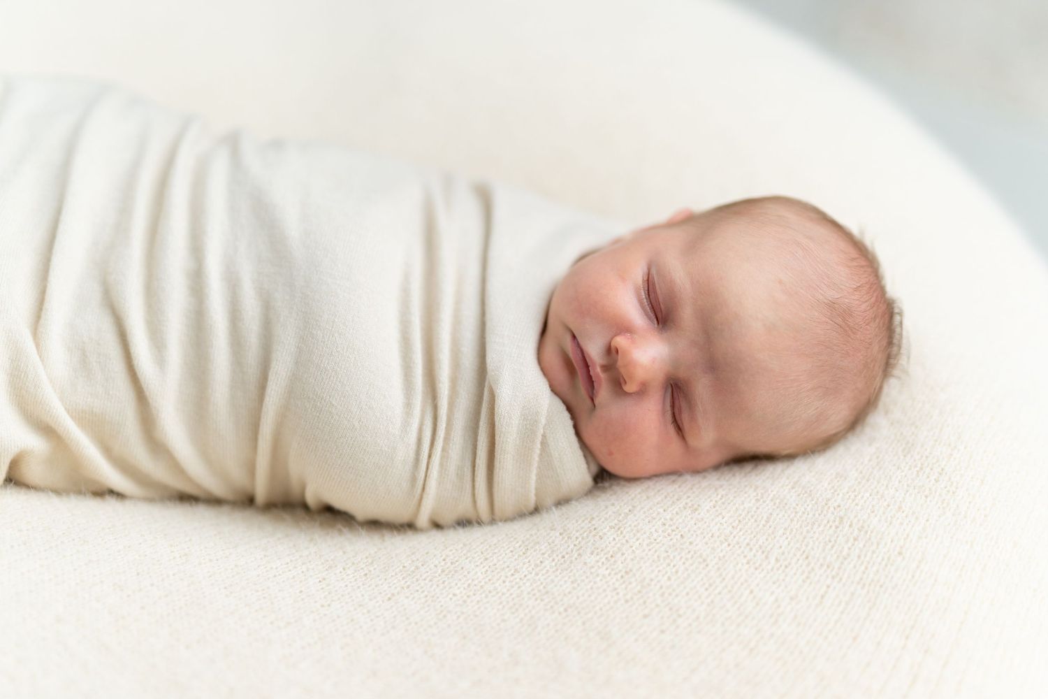 Peaceful newborn baby swaddled in a cream white wrap, sleeping soundly on a soft white blanket.