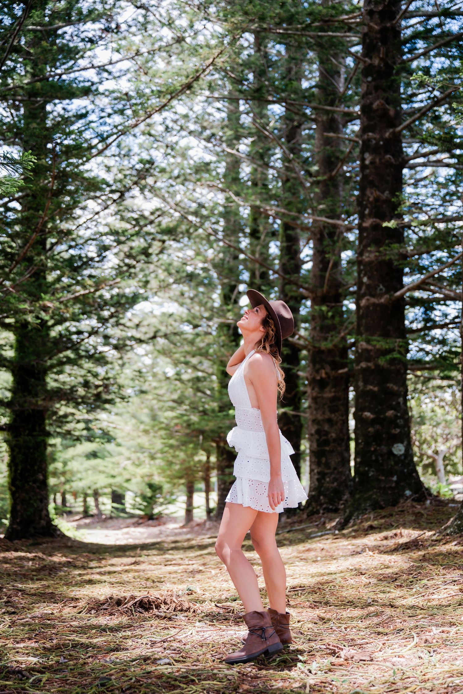 Woman in white dress and boots walks through a sunlit pine forest trail on a summer day.