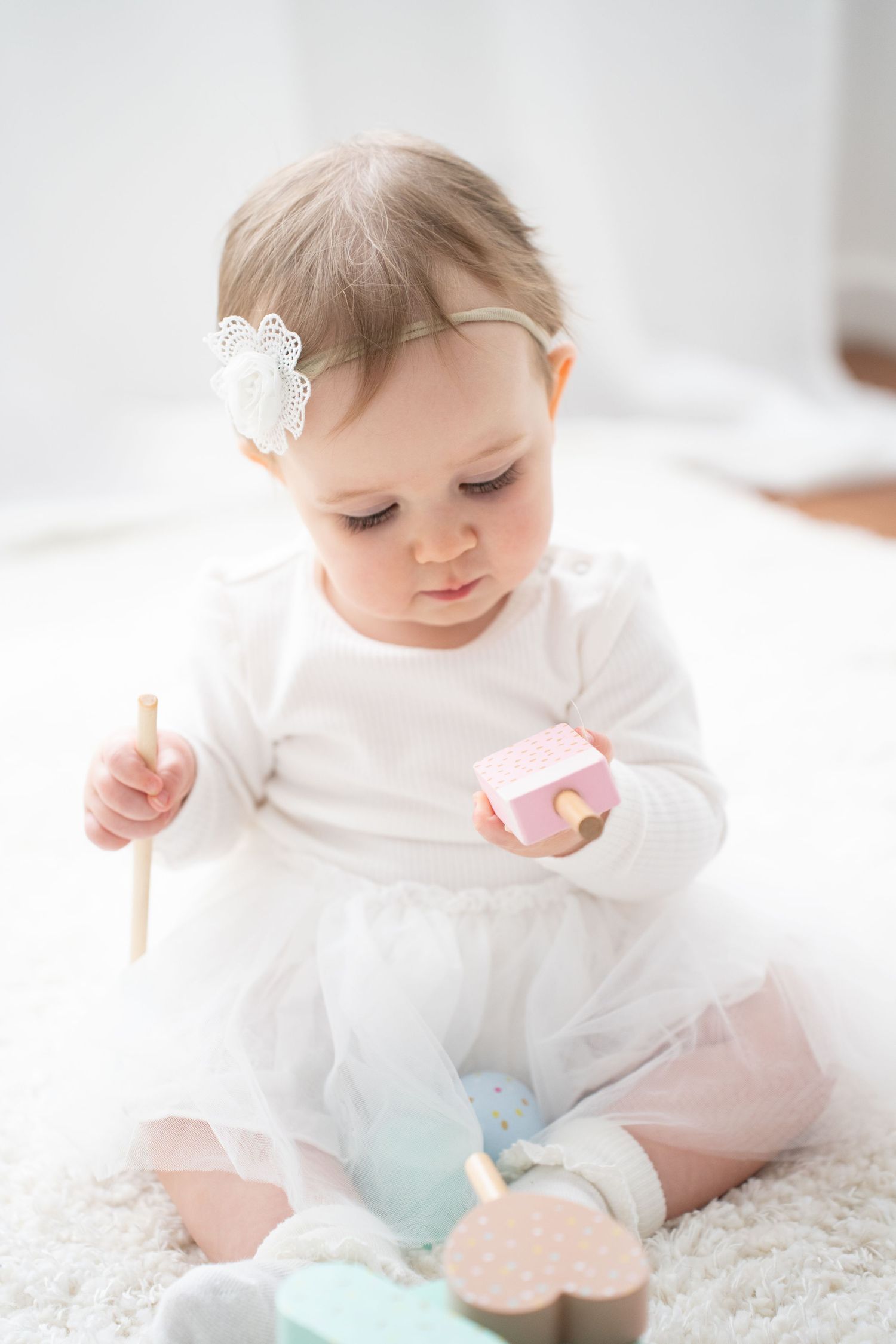 Adorable baby girl in white dress and floral headband playing with colorful wooden toys on a soft white carpet.