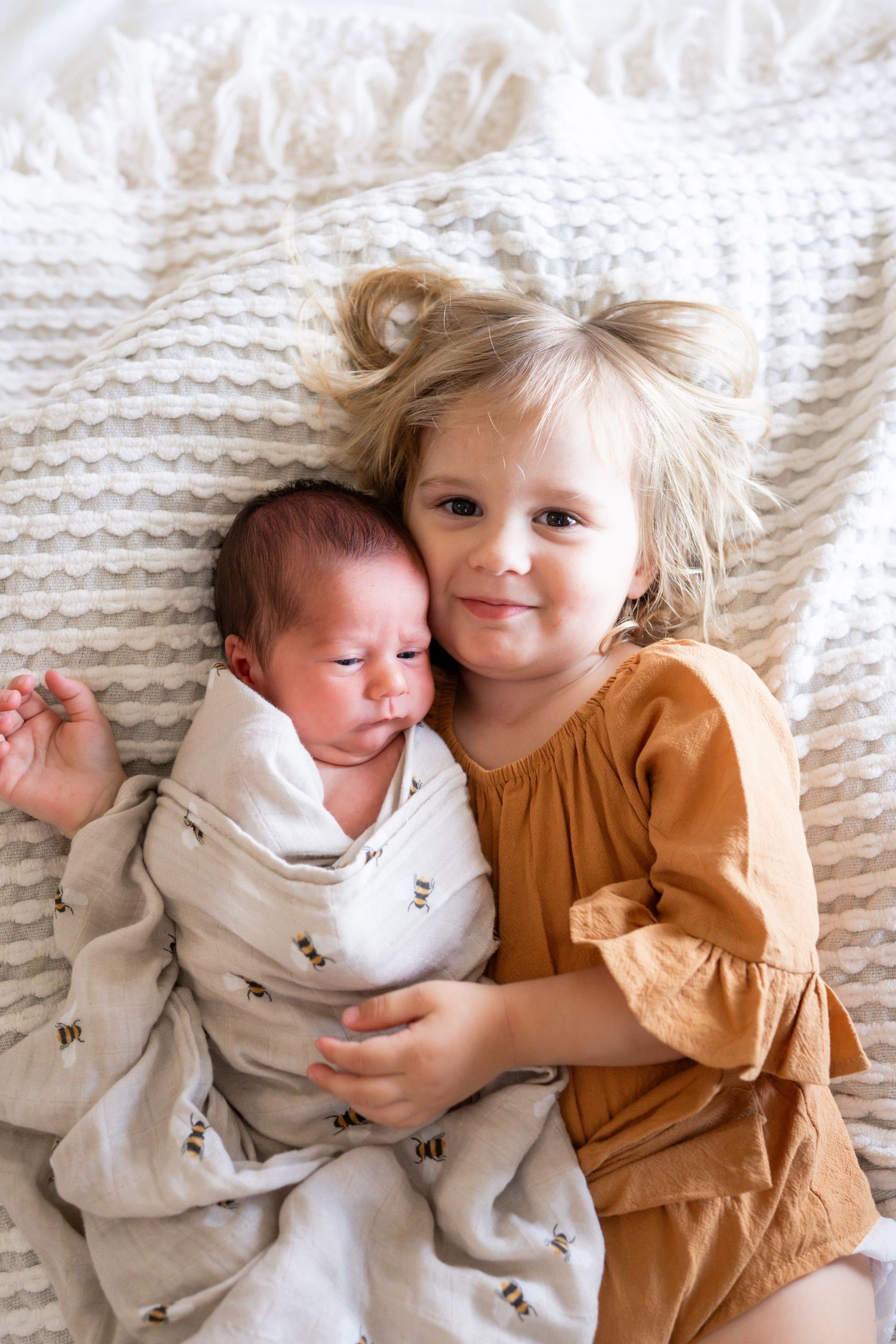 Toddler girl in rust dress smiles while holding swaddled newborn baby sibling on white textured blanket.
