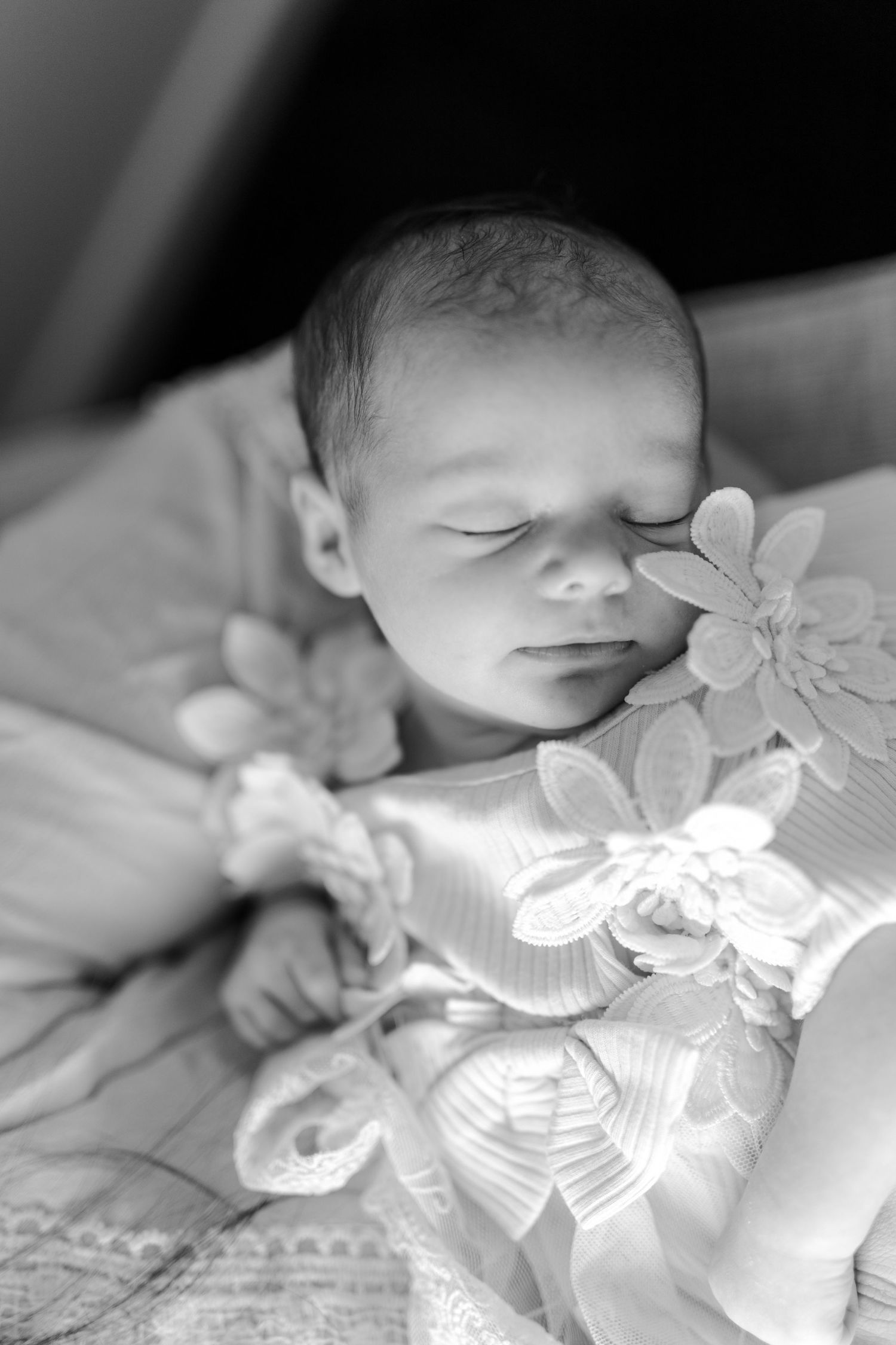 Black and white photo of a sleeping newborn baby girl in a floral dress with flower decorations near her face.
