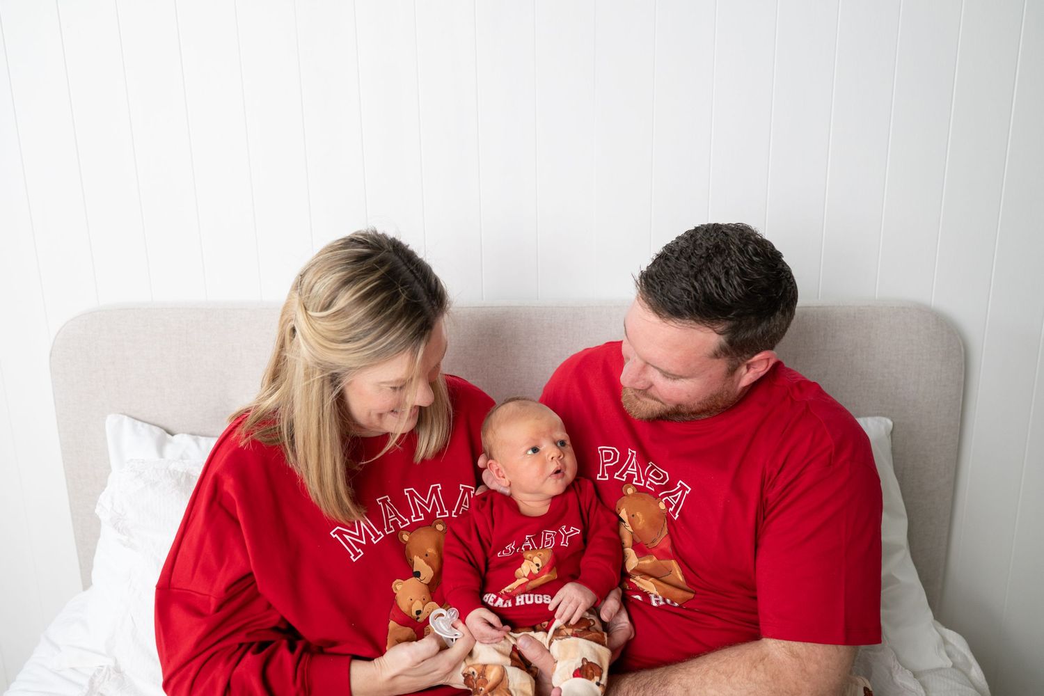 Smiling parents in matching red MAMA and PAPA shirts sit on a bed holding their baby, who wears a matching red outfit.