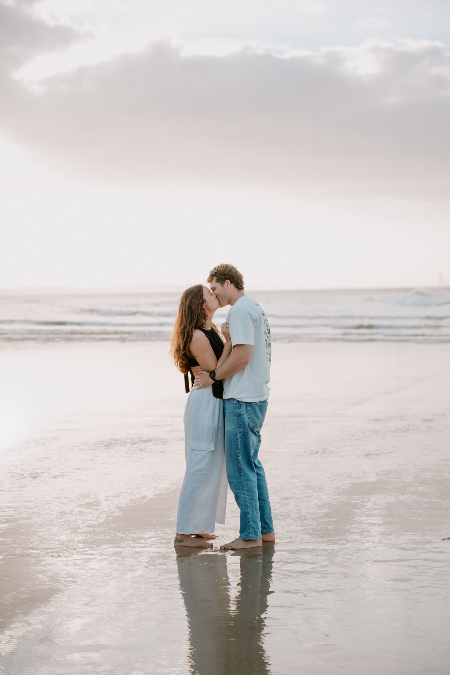 Couple sharing a romantic kiss on a reflective beach at sunset, woman in light blue pants and man in jeans.