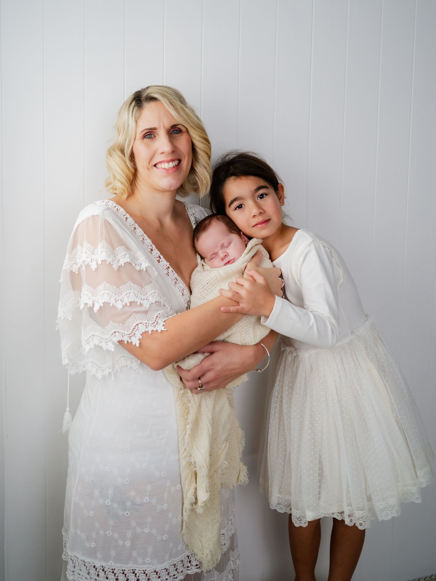 A smiling blonde woman in white lace dress holds a newborn while a young girl in white dress stands beside them.