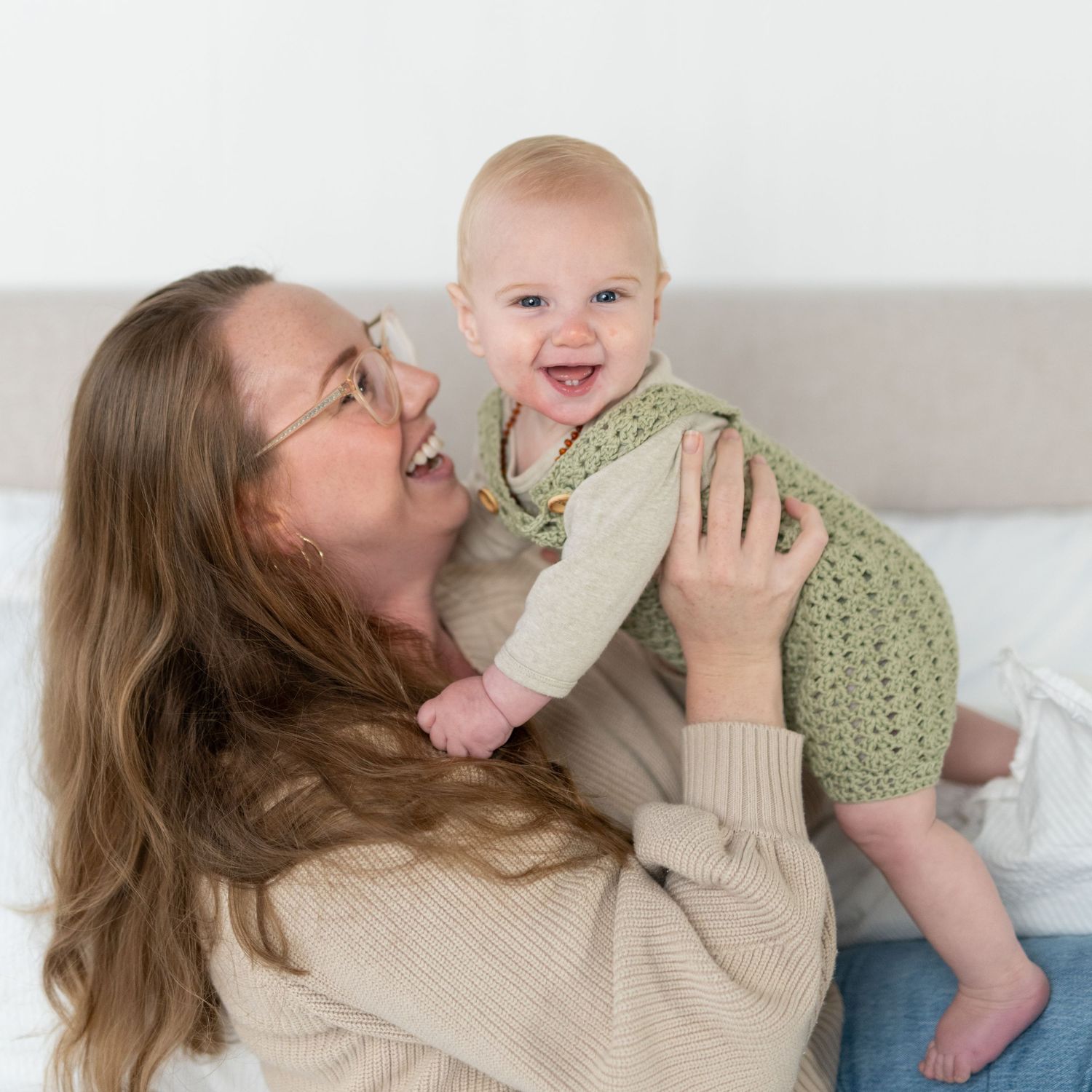 A smiling mother holds up her happy baby dressed in a green crochet outfit while sitting on a bed.