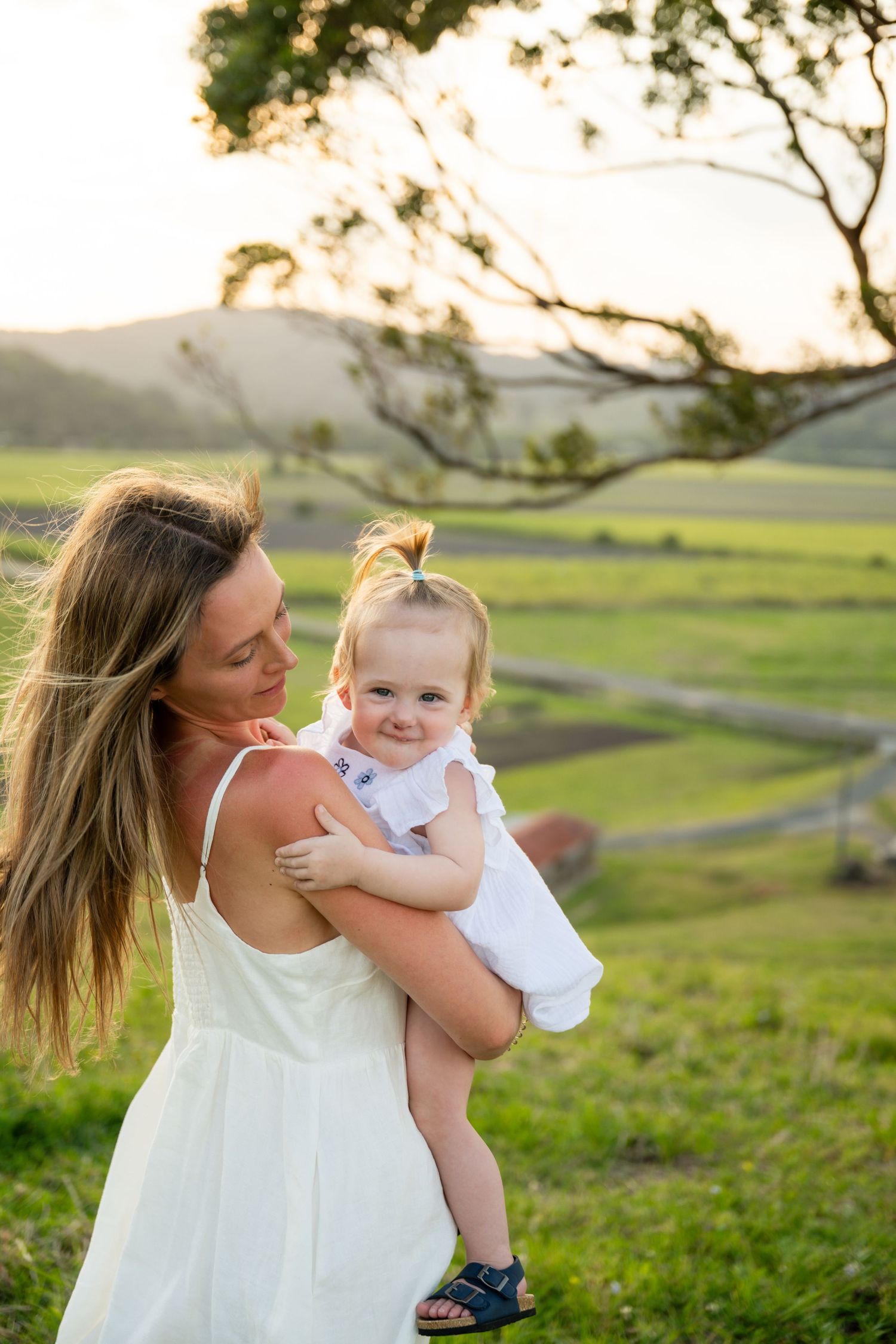 Parent and child in white dresses share tender moments while playing outdoors in a green meadow at sunset.