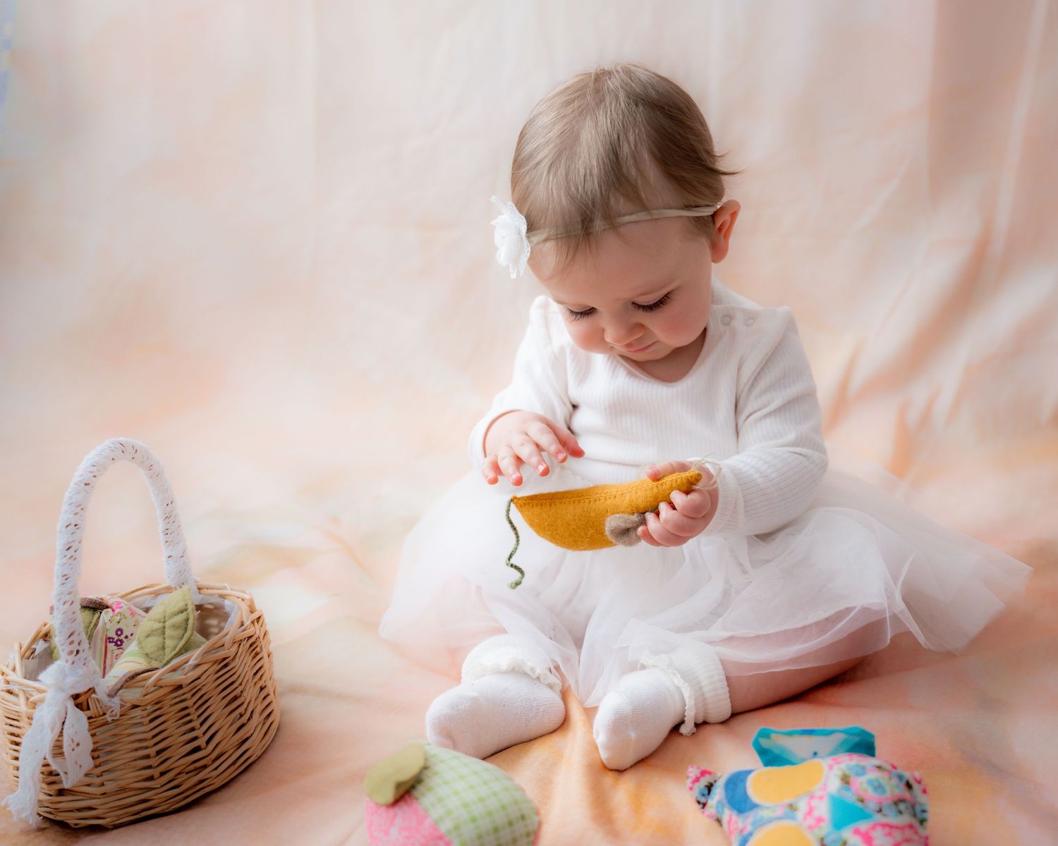 Baby girl in white dress plays with yellow toy banana near wicker Easter basket on soft pink background.