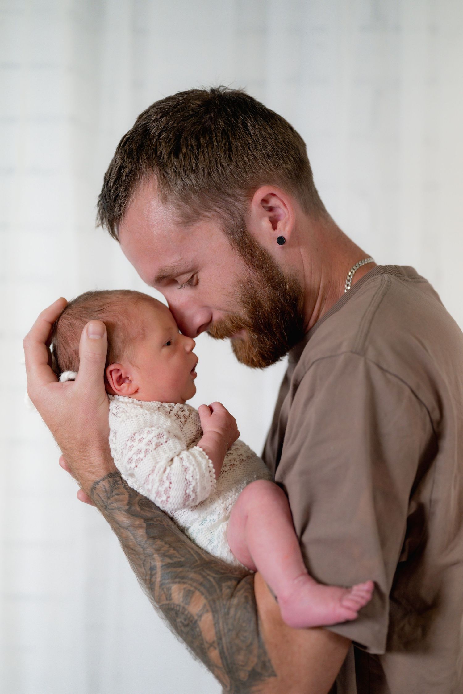 Bearded tattooed father tenderly touching noses with his newborn baby dressed in white lace, sharing a precious moment.
