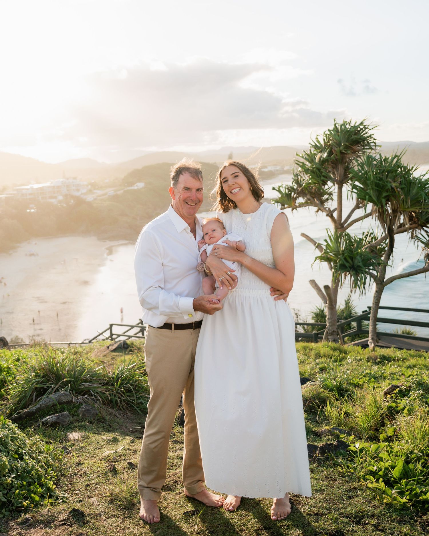 A happy family of three poses at a scenic coastal lookout during golden hour, parents holding their newborn baby.