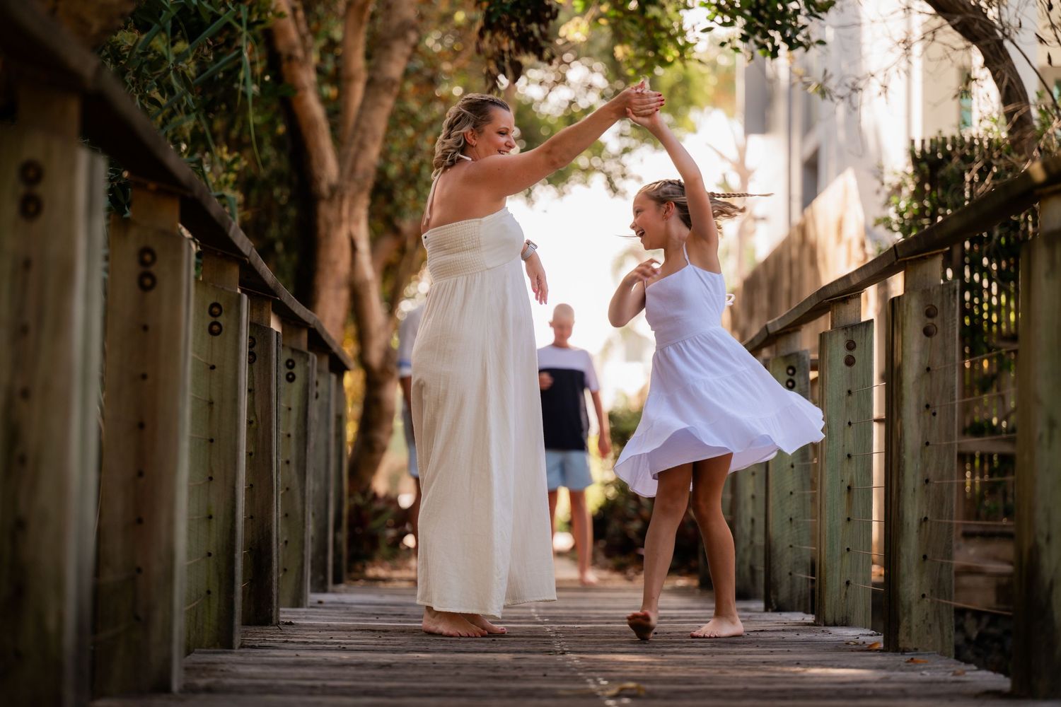 Three dancers in white dresses perform graceful movements in an outdoor corridor with stone columns and trees.