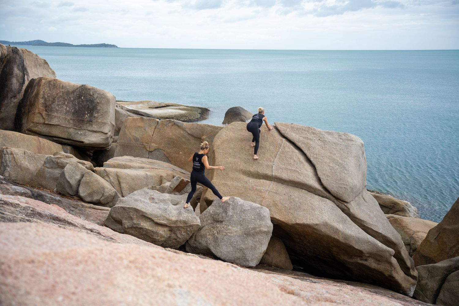 Two hikers climbing over large granite boulders along a scenic coastal trail with calm blue ocean in the background.