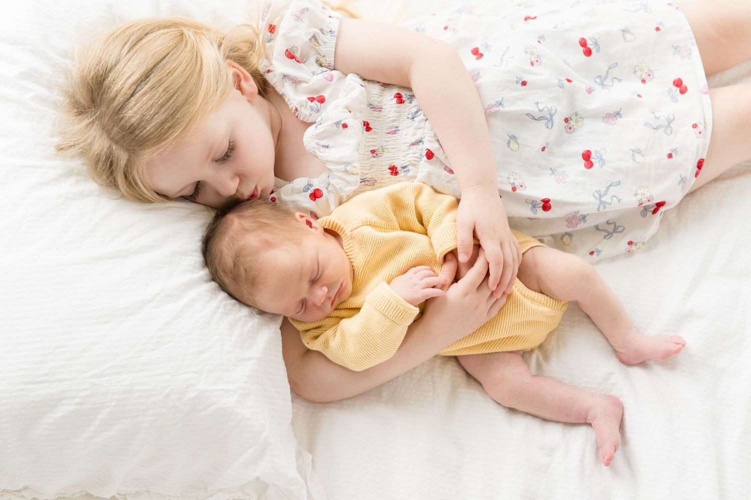 Young girl in floral dress lying on white bed, gently kissing sleeping newborn baby in yellow onesie.