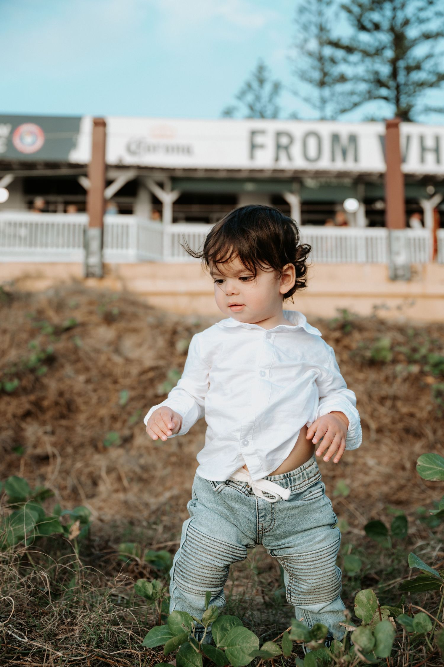 Toddler in white shirt and jeans standing outdoors near a building with a Corona sign.