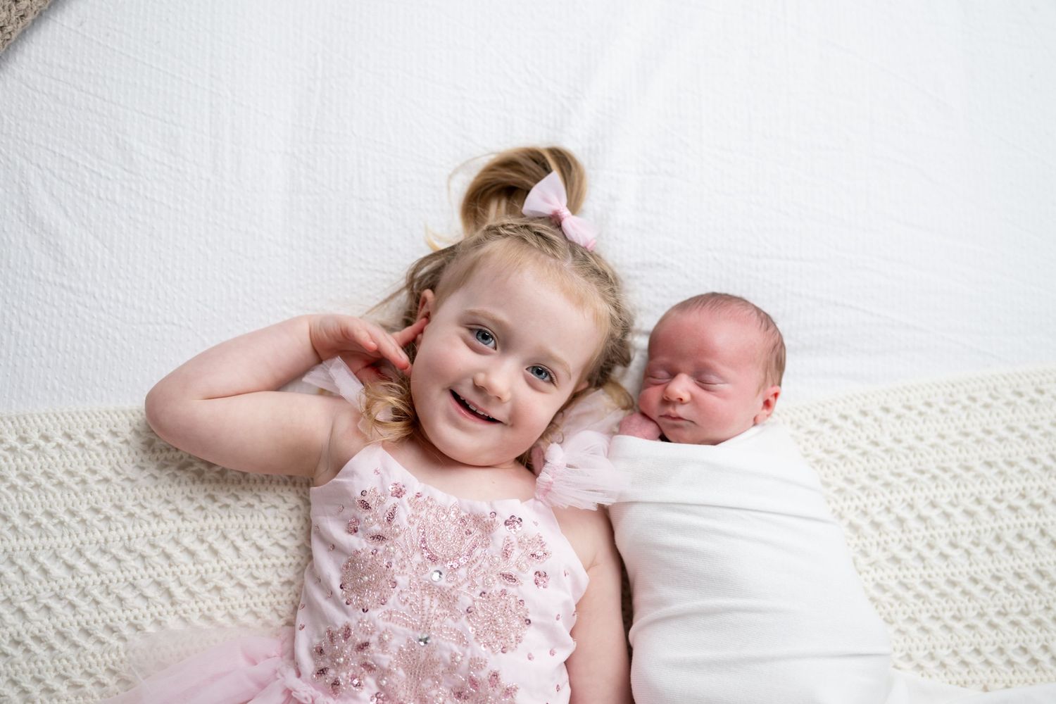 Smiling toddler girl in pink dress poses with sleeping newborn baby sibling on white knit blanket.