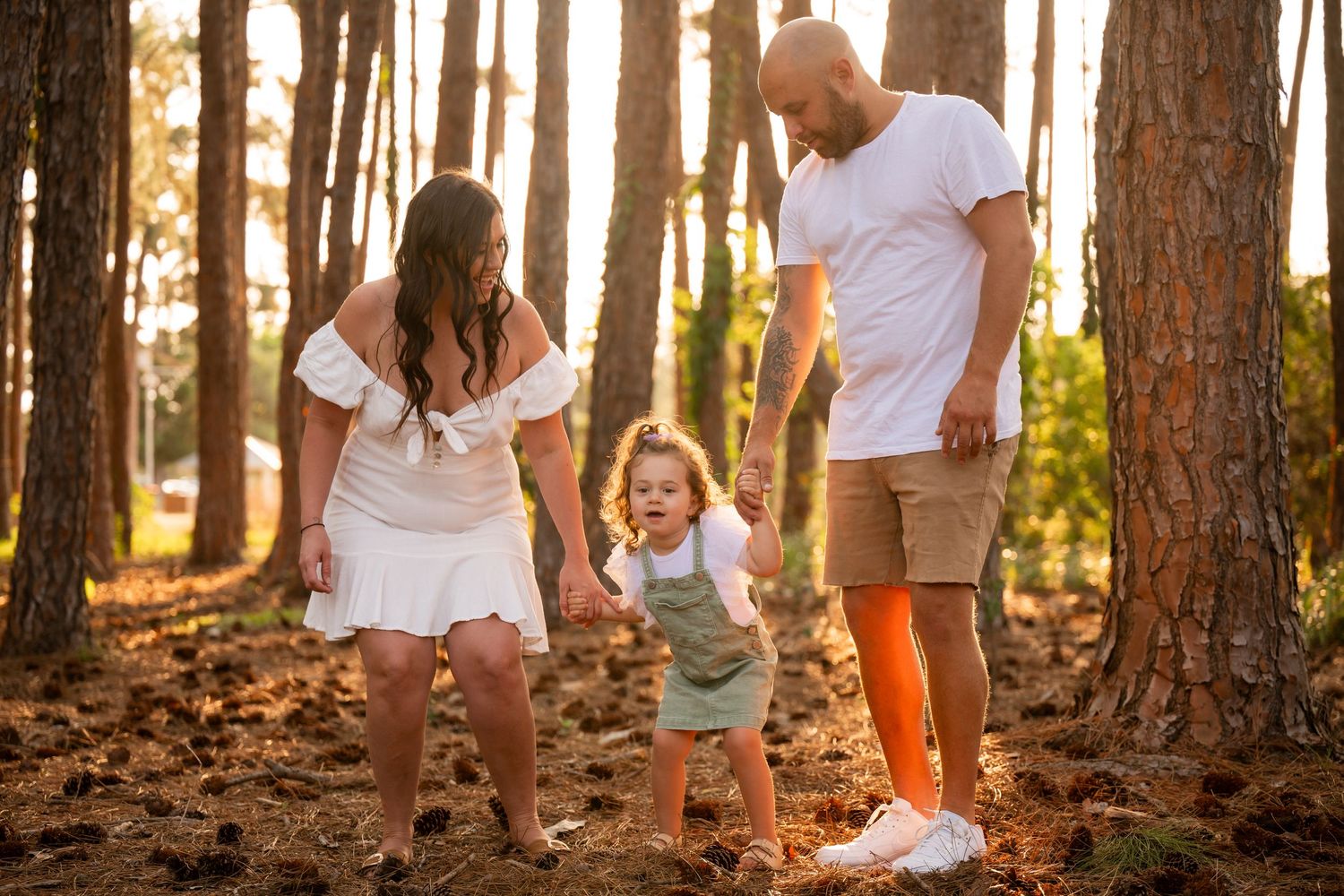 Family of three enjoying a golden hour stroll among tall pine trees, parents watching toddler walk in forest.