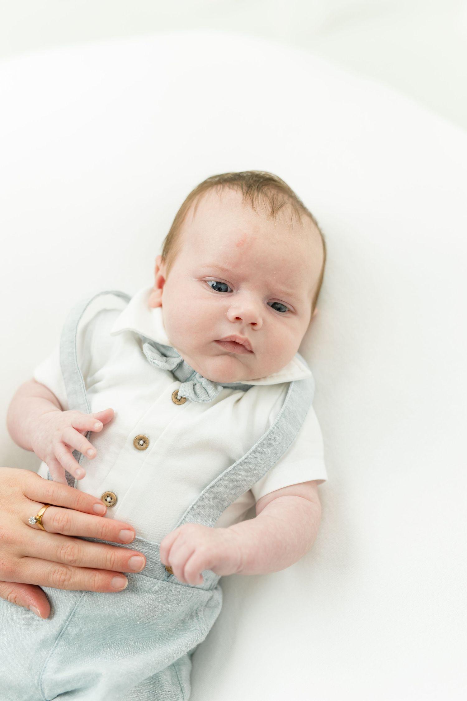 Newborn baby in white onesie and gray overalls lying on white background with parent's hand visible.
