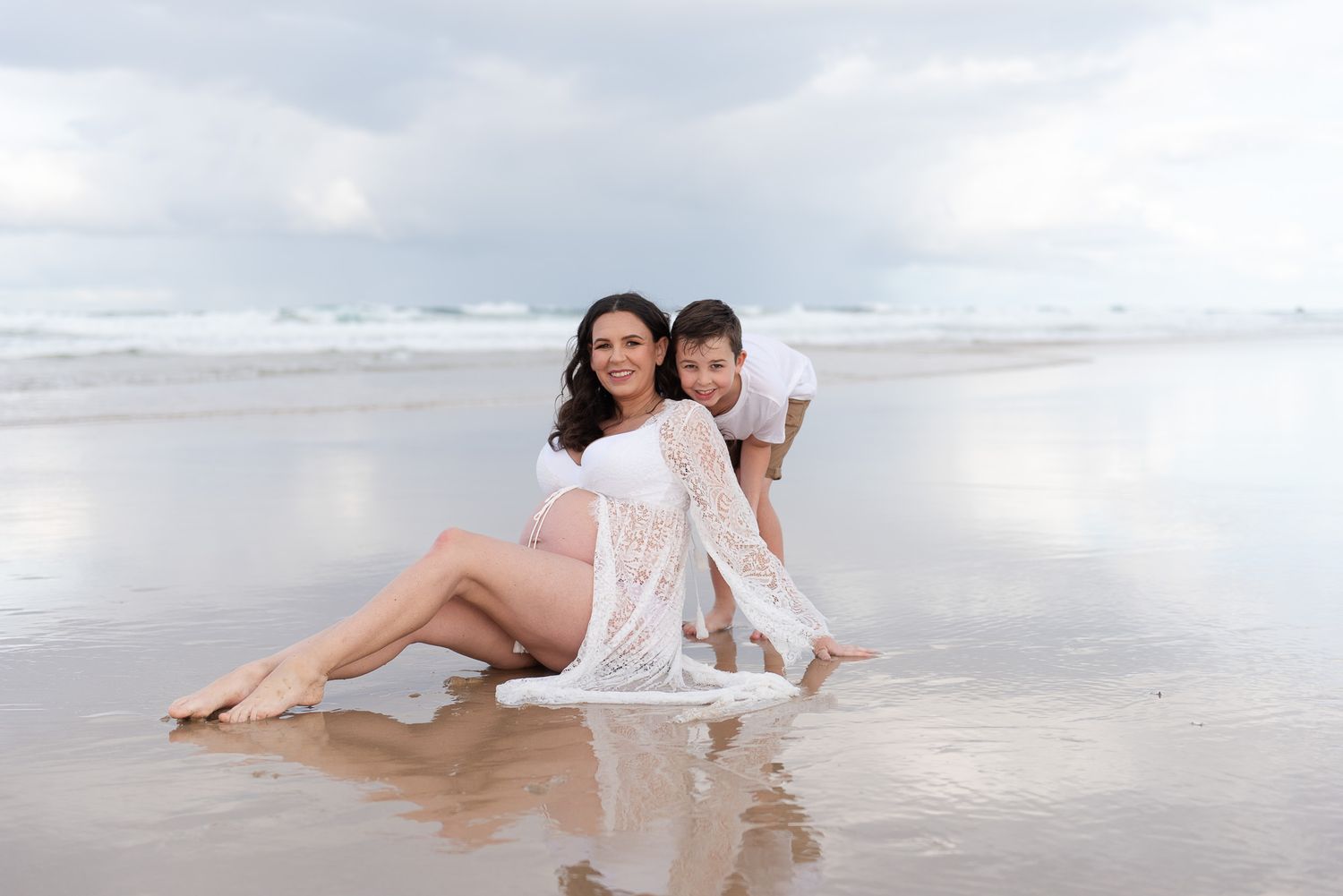 Pregnant woman in white lace dress sits on beach with partner, ocean waves reflecting on wet sand.