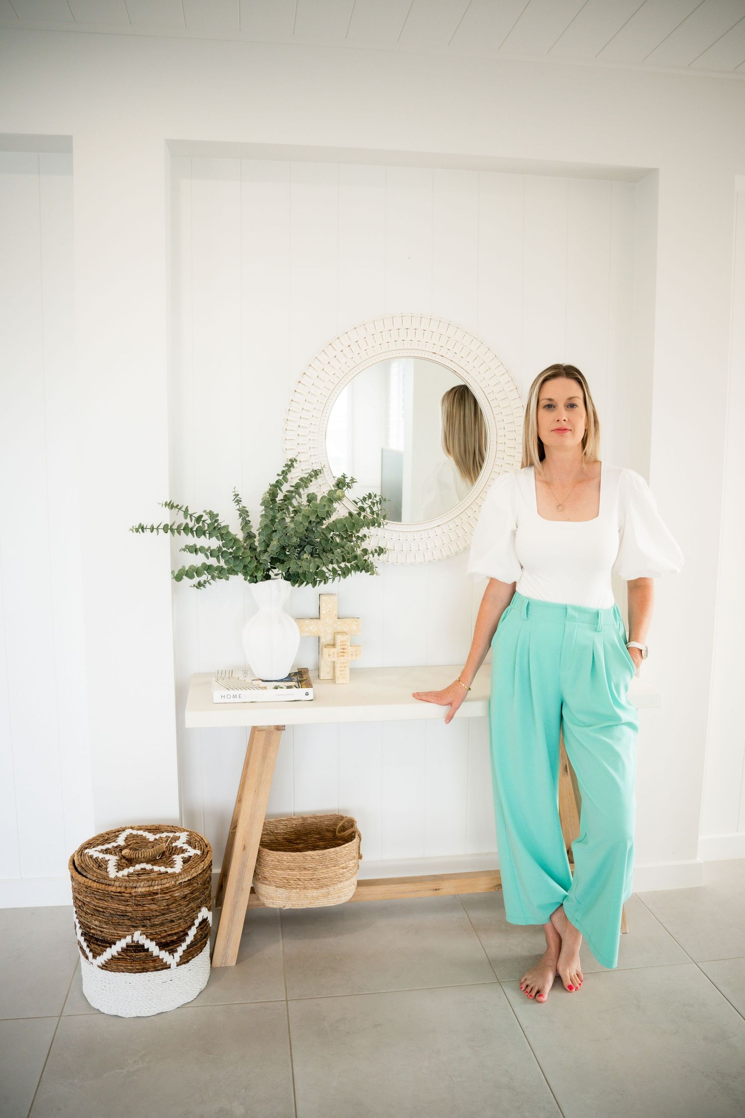 A person wearing a white t-shirt and mint green pants stands in a bright, minimalist room with potted plants and woven baskets.