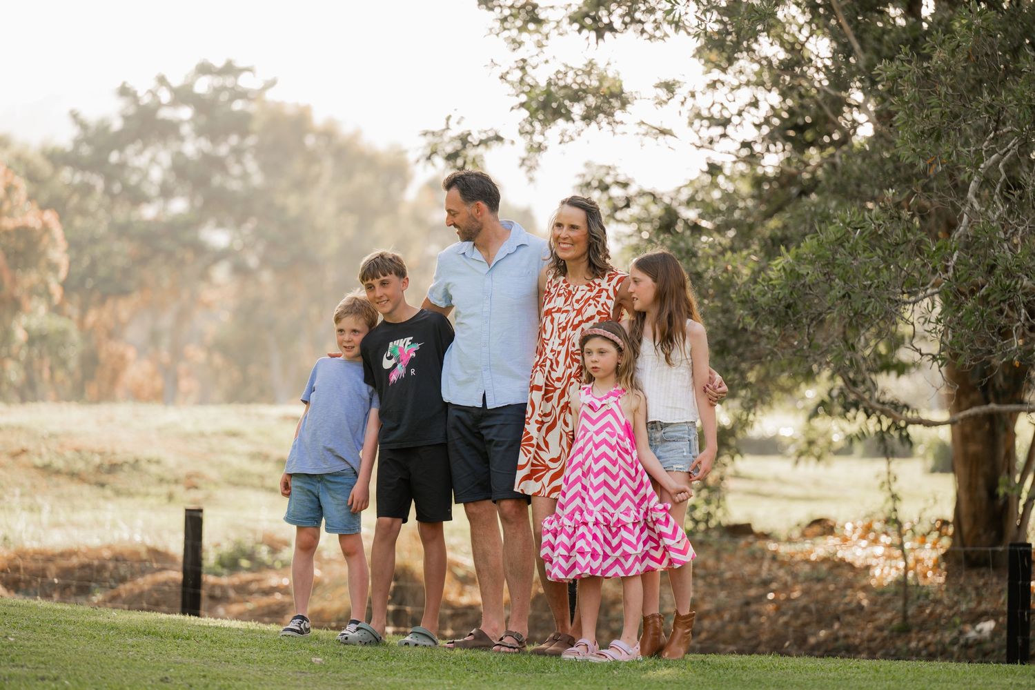 Family of six standing together on a green lawn in a sunlit park, smiling and embracing during a golden hour outdoor portrait.