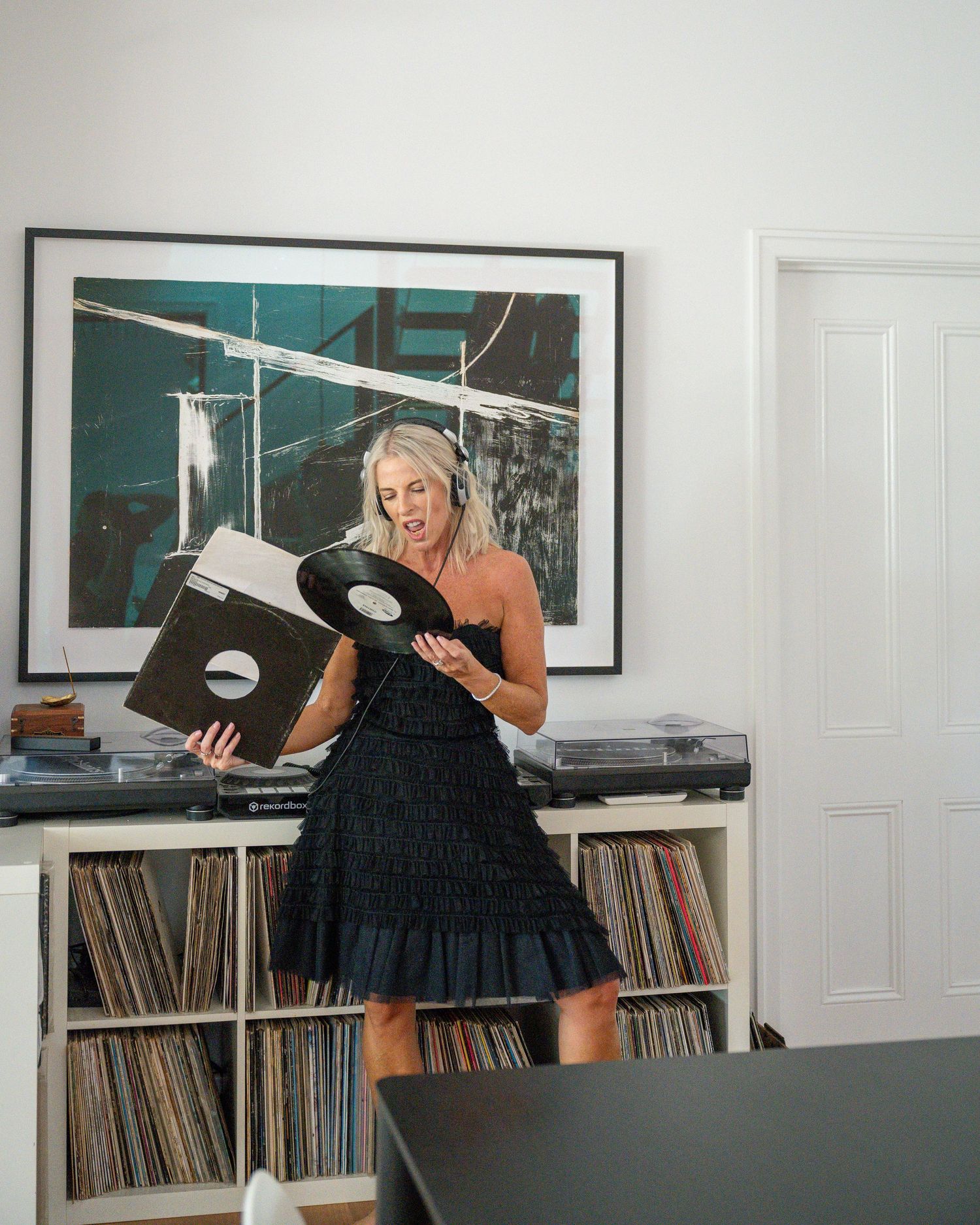 Woman in black dress holding vinyl record near shelves of records with turntable and abstract artwork on wall.