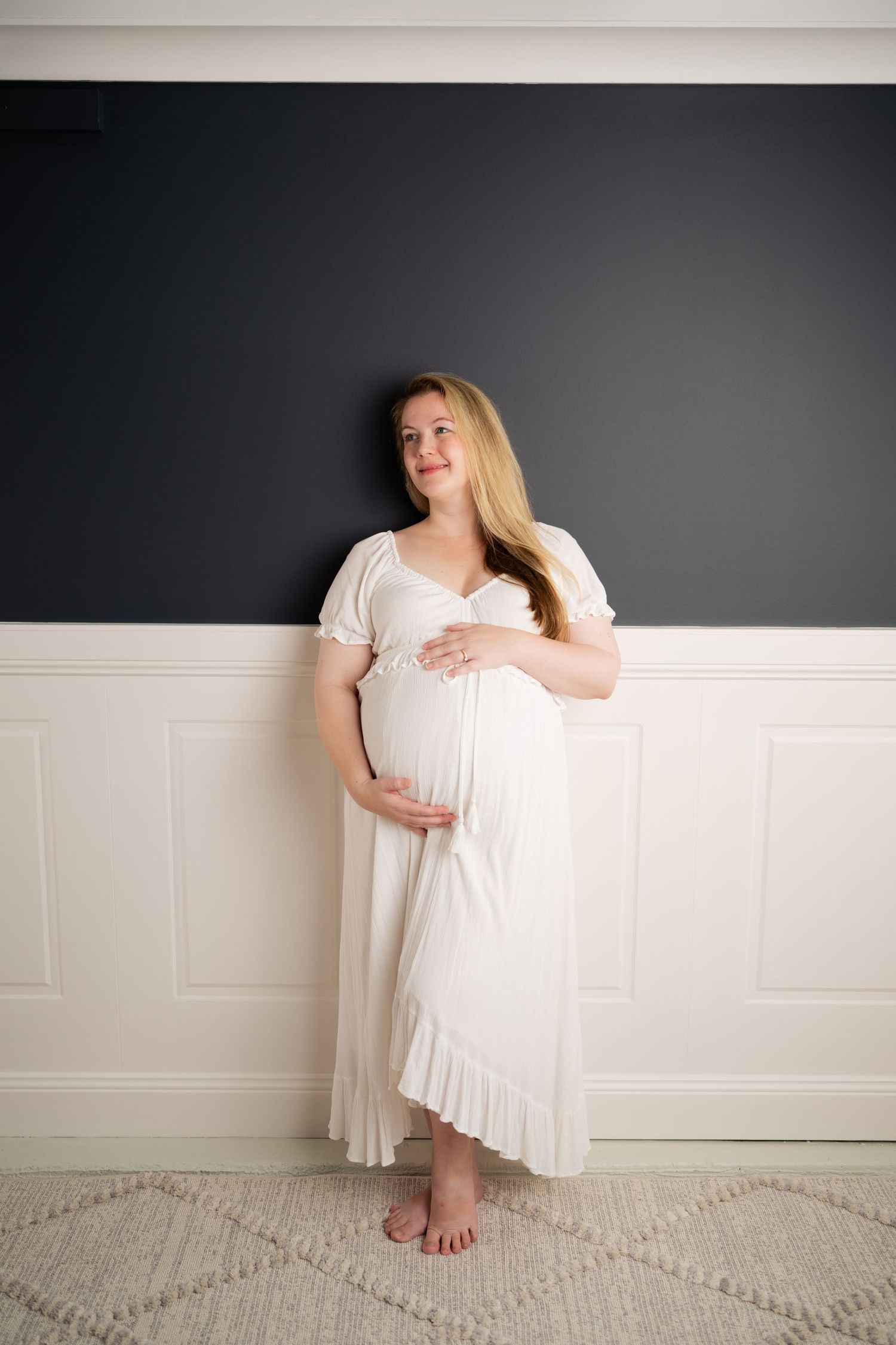 Pregnant woman in white dress cradling baby bump, leaning against dark wall, maternity photography.