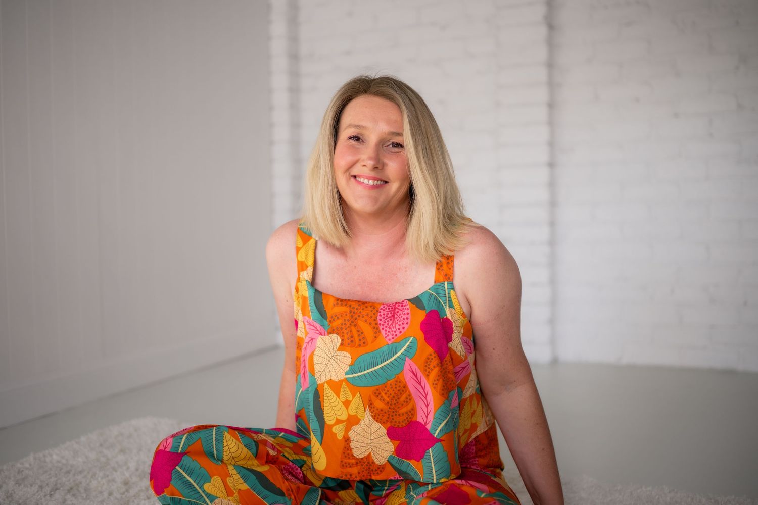 Woman smiling in colorful floral jumpsuit seated against white brick wall.