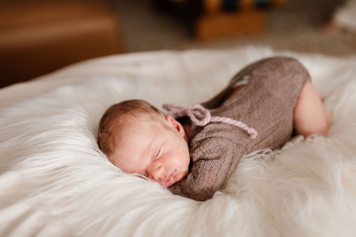 Newborn baby sleeping peacefully on white fluffy blanket wearing brown knit romper and bonnet.