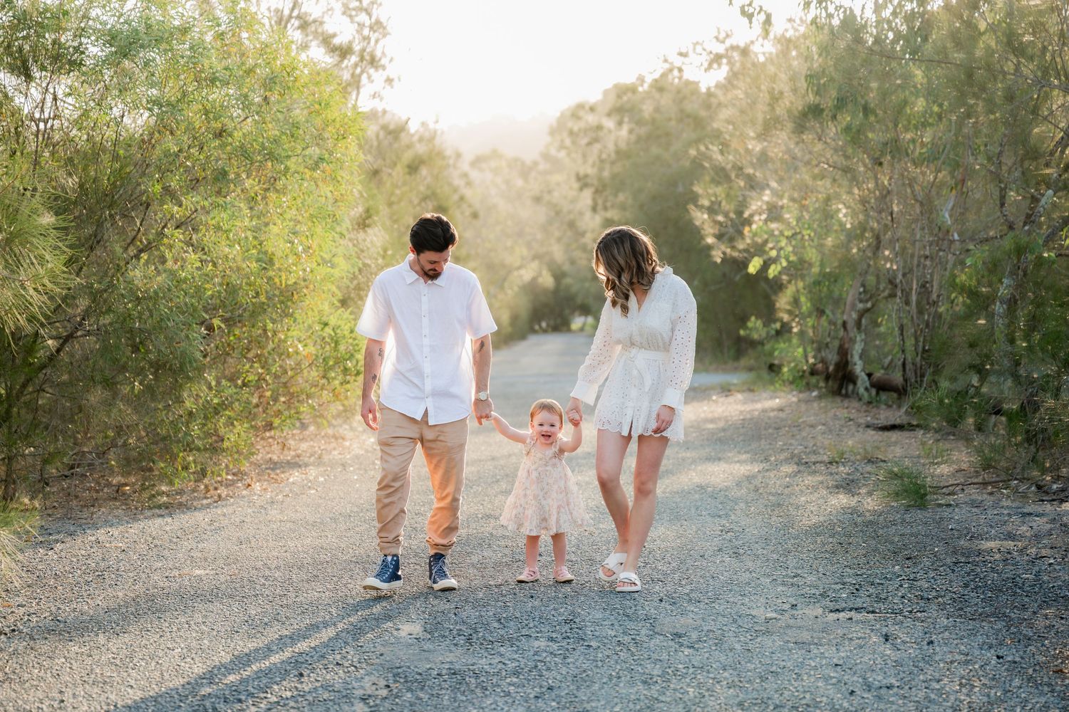 Family of three walking on a gravel path, parents holding toddler's hands outdoors.
