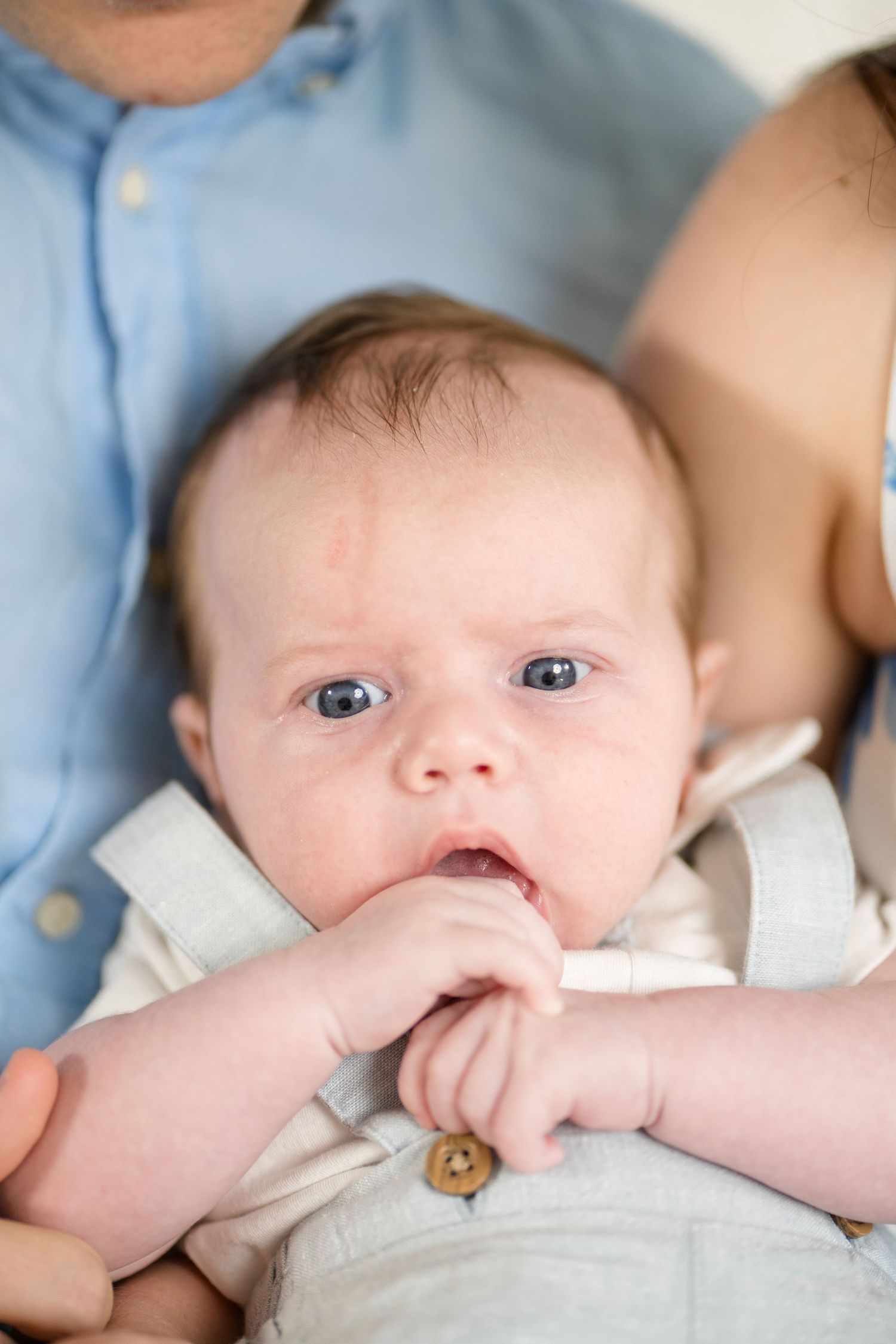 Adorable newborn baby with blue eyes sucking fingers, cradled in parents arms, wearing a cream romper.