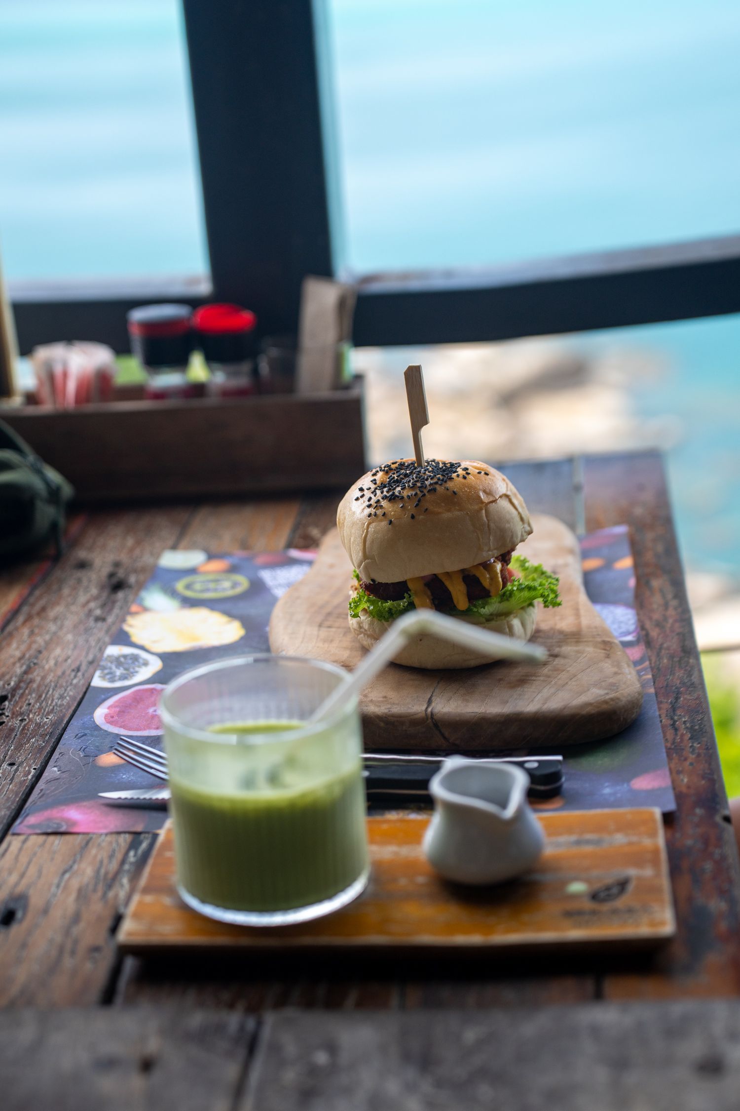 Gourmet burger on wooden board with green smoothie, served at oceanview restaurant table.